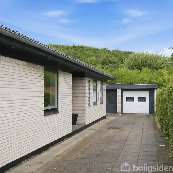 Et hvidt murstenshus står stille ved en flisebelagt indkørsel. Omgivet af tæt, grøn beplantning ses en garage i baggrunden under en blå himmel.