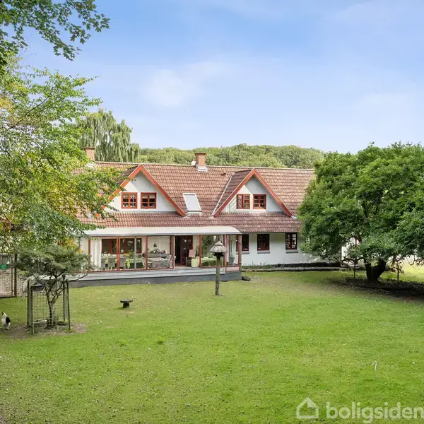 En stor villa med rødt tag står midt i en grøn have omgivet af træer og en flagstang til højre. Huset har flere vinduer og en veranda.