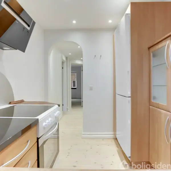 En moderne køkkenindretning viser en el-komfur med emhætte til venstre, træskabe til højre, og en lys gang, der fører til andre rum i en bolig med hvidmalede vægge.