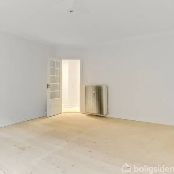 Et tomt rum med lys træbund, en åben dør til en oplyst gang og en radiator på væggen. Væggene er hvide, og der er ingen møbler.