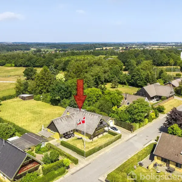 Hus markeret med en rød pil, omkranset af grønne områder og naboer. En dansk flagstang står i forhaven. Byhuse og marker kan ses i baggrunden.