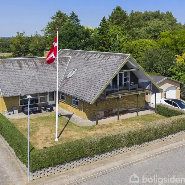 Et gult murstenshus med skrånende tag og en balkon står i en grøn have. En flagstang med Dannebrog står foran huset. En hvid bil er parkeret i indkørslen.