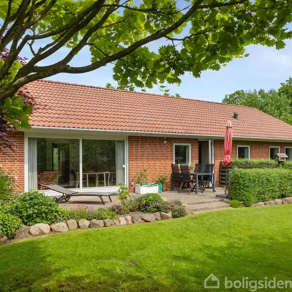 Et rødt murstenshus med rødt tegltag står i en frodig have. Der er et terrassemøblement og parasol på en flisebelagt terrasse foran huset, omgivet af grønne buske og træer.