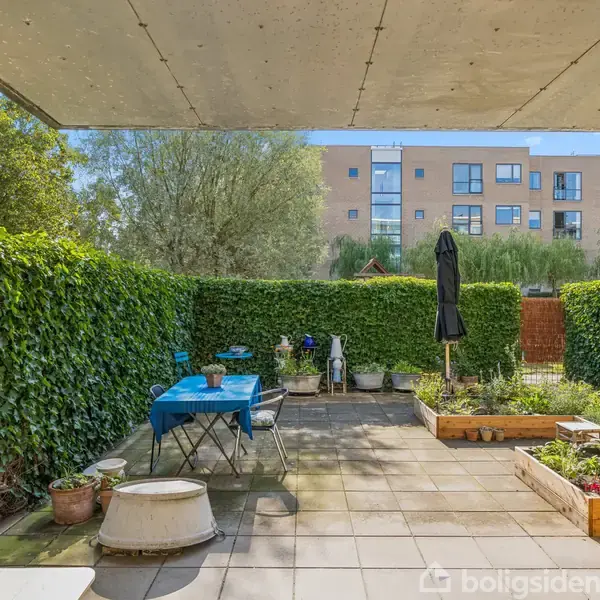 Terrasse med borde og stole står på flisebelagt gulv, omringet af grønne hække og plantekasser. En parasol er opstillet, og i baggrunden ses en murstensbygning under en blå himmel.