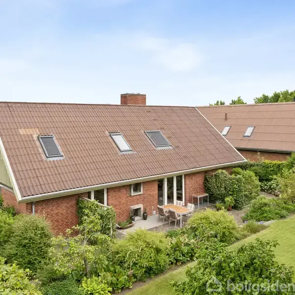 Hus med rødt murstensfacade står stille omringet af en velplejet græsplæne og tæt grøn bevoksning, under en klar blå himmel.