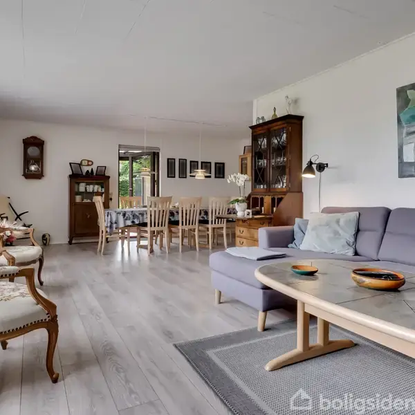 En stue med en blå sofa og sofabord, dekoreret med skåle, i et indrettet rum med spisebord og stole i baggrunden. Væggene har kunstværker og et vitrineskab.