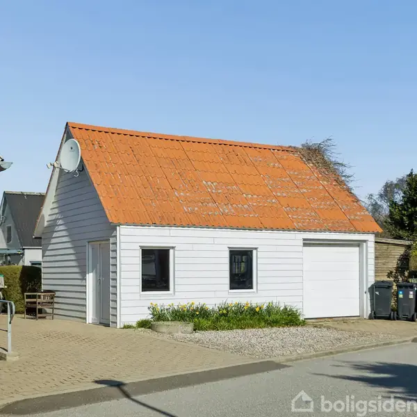 Hvid garagebygning med orange tag og en parabolantenne, står ved fortovet i et villakvarter. Omkringliggende huse og træer ses i baggrunden under en blå himmel.