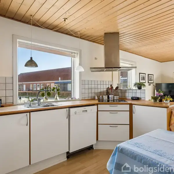 Køkken med hvide skabe og træbordplade, indbygget opvaskemaskine og kogeplade. Store vinduer giver naturligt lys. Spisebord med blå dug i forgrunden. Moderne og lys atmosfære.