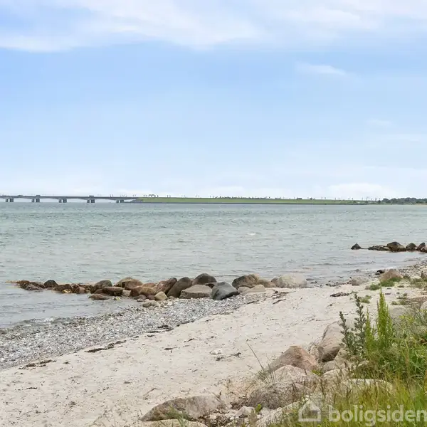 Strand med sand og sten, roligt vand i bevægelse, med en bro i baggrunden. Græs og buske vokser langs kysten.