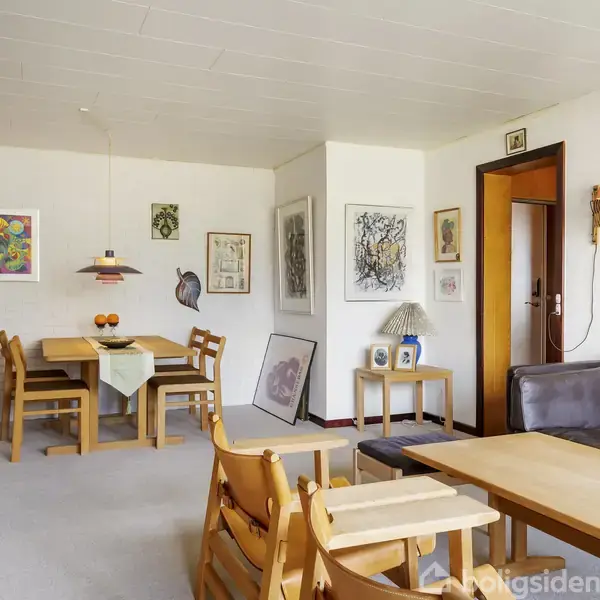 Stue med borde og stole, der skaber en hyggelig spisestueatmosfære. Væggene er dekoreret med kunst, og en lampe hænger over spisebordet. Sofaen står ved siden af en dør.