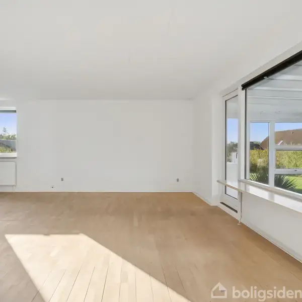 Et tomt rum med trægulv, store vinduer der lukker lys ind, og udsigt til en have udenfor. Væggene er hvide, og rummet har en lys og åben atmosfære.