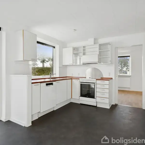 Et moderne køkken med hvide skabe står tomt og velordnet. Der er en indbygget ovn, to store vinduer med udsigt og en glasdør fører ud til en terrasse.