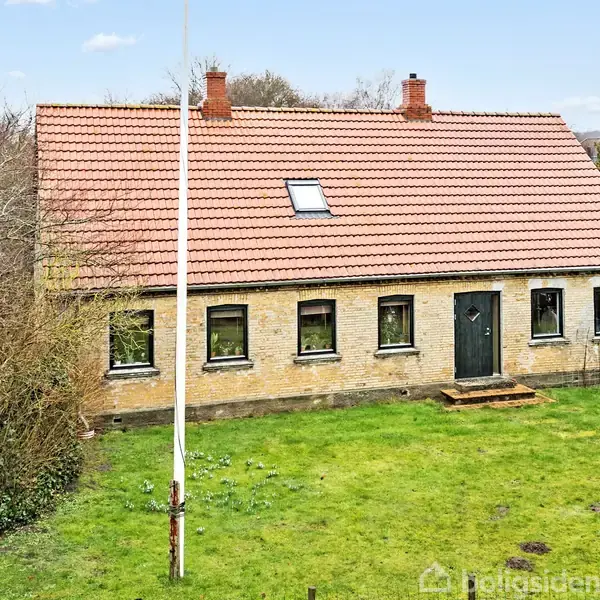 Et gult murstenshus med rødt tegltag står på en grøn græsplæne. Huset har seks vinduer, en mørk dør og en flagstang foran. Træer ses i baggrunden.