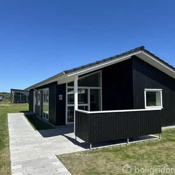 Sort træhus står stille på en grøn græsplæne under en skyfri blå himmel, omgivet af en flisebelagt sti. Der er ingen tekst i billedet.