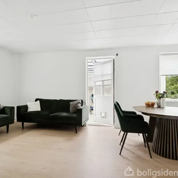 En stue med to mørkegrønne sofaer til venstre, et rundt spisebord med fire stole til højre, og en åben dør til en terrasse. Væggene er hvide.