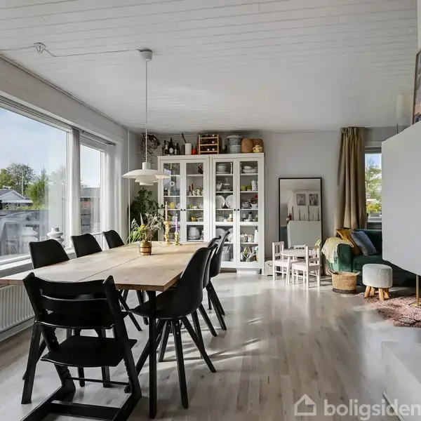 Et spisebord med sorte stole står i et lyst spiseområde, omgivet af store vinduer, hvidmalede vægge og et vitrineskab fyldt med genstande. En lampe hænger over bordet.