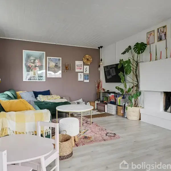 Stue med grøn sofa prydet med farverige puder, ved siden af en åben pejs med planter og stearinlys på toppen. Væggene er dekoreret med billeder, og et børnebord står forrest.