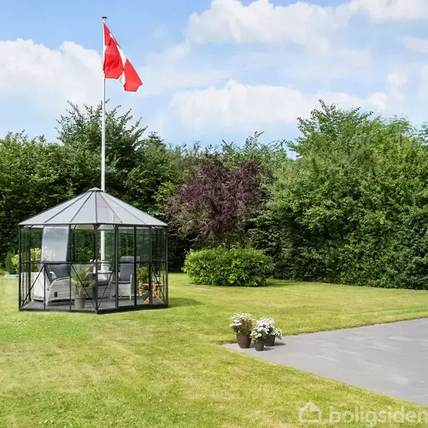 Glaspavillon står på en græsplæne med flagstang i nærheden, omkranset af buske og træer. En terrasse ses til højre med blomsterdekorationer.