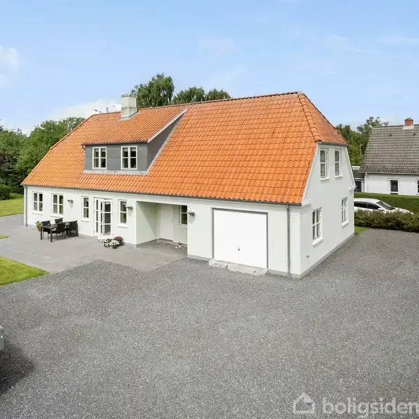 Et hvidt hus med orange tag står stille på en grusbelagt plads ved siden af en velholdt græsplæne. En flagstang og drivhus ses i baggrunden.