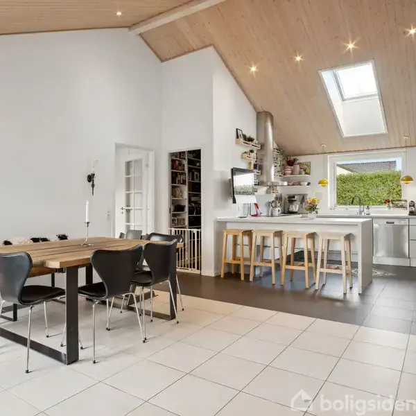 Et moderne køkken-alrum med spisebord og sorte stole i forgrunden. Bagved er en køkkenø med barstole under et skråt, lysindfaldsrigt loft med ovenlysvindue. Indretning er lys og rummelig.