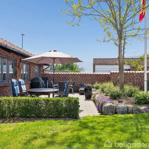 Terrasse med havemøbler står under en parasol ved siden af et murstenshus. En græsplæne omgiver området, og et lille træ med en flagstang står til højre. Klar himmel.