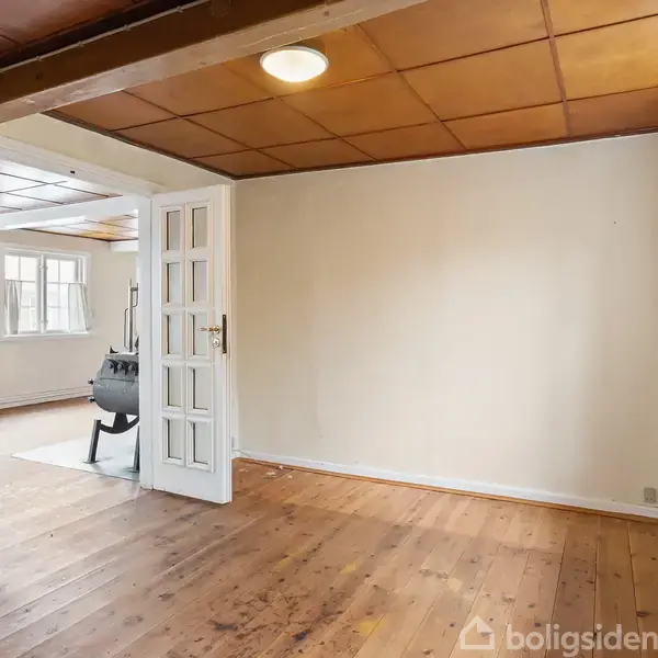 Et tomt rum med trægulv og træloft, adskilt af en dør til en stue med brændeovn. Sollys strømmer ind gennem vinduer i et lyst og enkelt interiør.