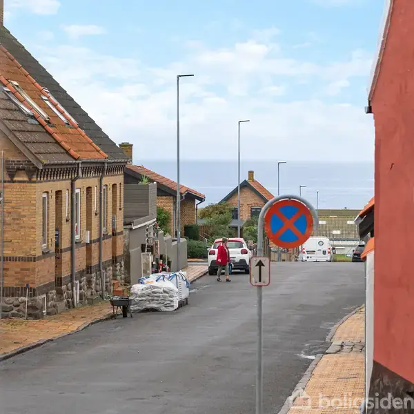 Gade, der fører ned mod havet med biler parkeret langs vejen. Huse på begge sider, et skilt for parkering forbudt midt på gaden, under en delvist skyet himmel.