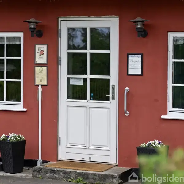 Hvid dør med glasruder til venstre for to hvide vinduer i en rød facade. To sorte lanterner hænger over døren. To blomsterkrukker står ved siden af døren. Tekst: “Hinds Hotel” og “Plumploe”.