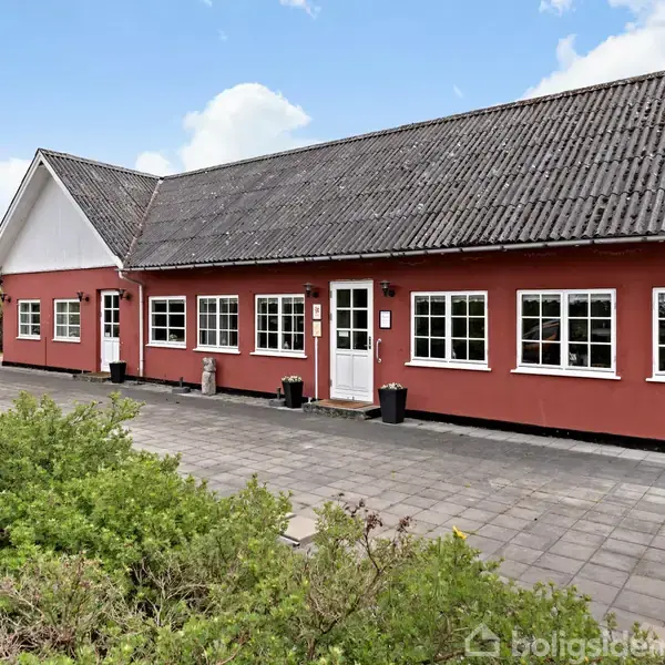Rødt enplanshus med sort tag, flere vinduer og en hvid dør. Skilt til venstre med teksten: "Restaurant Gaarden Selskabslokaler". Flisebelagt forplads og buske omkranser bygningen.