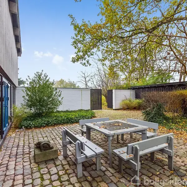 Gråt bord og bænke på en brostensbelagt terrasse med omgivelser af træer og buske. Et moderne hus ses til venstre med vinduer, og der er en hvid mur i baggrunden.