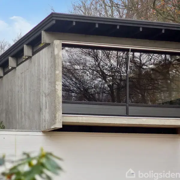 Et moderne træhus med store vinduespartier reflekterer træerne udenfor. Huset står i et skovområde med en klar himmel i baggrunden.