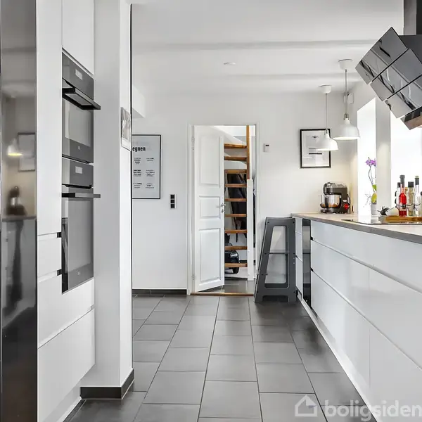 Et moderne køkken med hvide skabe og indbyggede apparater. Lang bordplade til højre med barstol. Sorte fliser på gulvet og et åbent rum, der leder til en trappeopgang i baggrunden.