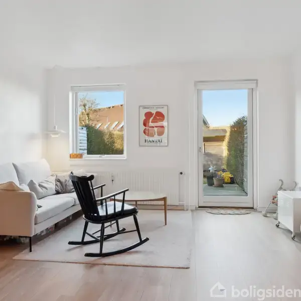 Stue med hvid sofa og sort gyngestol på tæppe. Tv hænger på væggen til højre. Store vinduer lukker lys ind, med udsigt til have. Plakattekst: "Tivoli 1975".