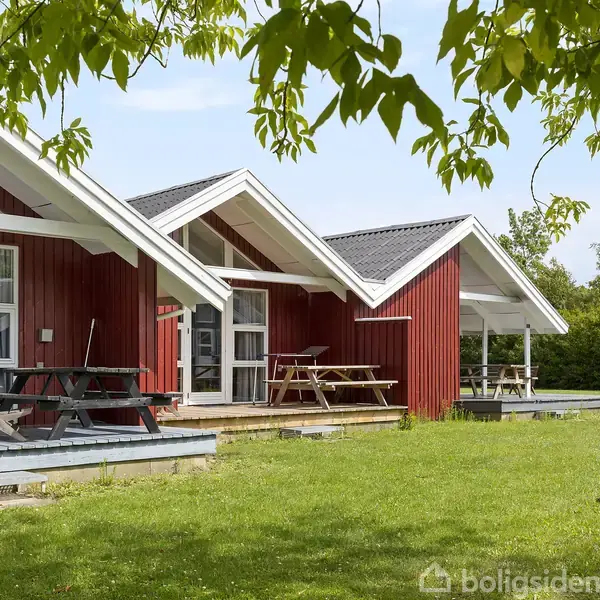 Rødt træhus med hvide tage står på en grøn græsplæne. Der er picnicborde foran huset, og træer omkranser omgivelserne, hvilket skaber en naturlig og rolig atmosfære.