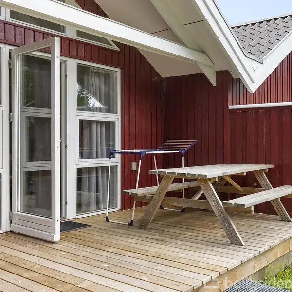 Et rødt træhus med en veranda. På verandaen står et træbord med bænke. Bagved ses en glasdør, solen skinner, og omgivelserne er grønne.