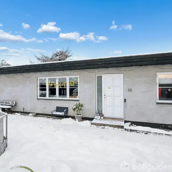 Et gråt, enkeltplanshus dækket af sne står i en vinterhave, med en sort tagkant og store vinduer. Husnummeret 22 ses ved siden af hoveddøren.