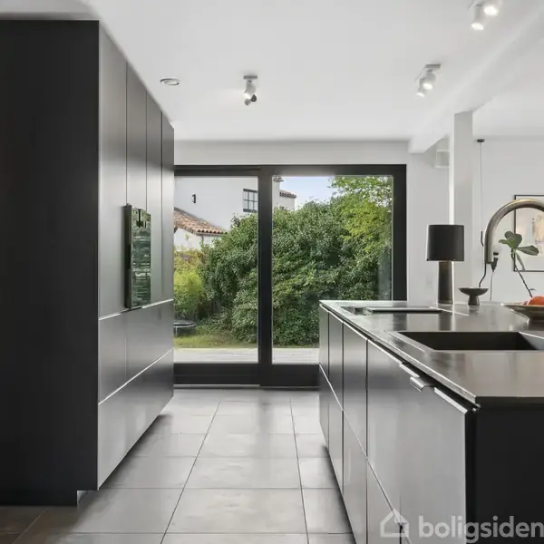 Køkken med mørke skabe og en central køkkenø, udstyret med en vask. Store vinduer åbner udsigten til en have med buske. Minimalistisk indretning, lyst og moderne design.