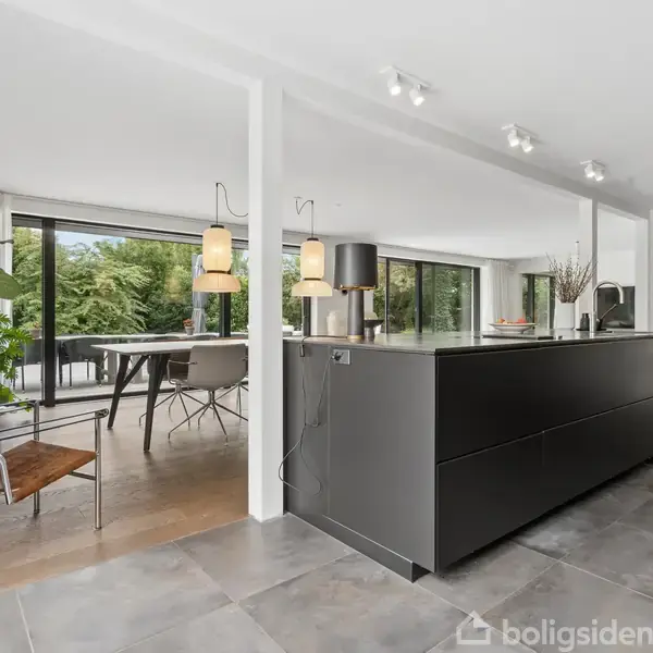 En moderne villa med store vinduer og en rummelig træterrasse fyldt med havemøbler. Omkring huset er der frodige, grønne træer, hvilket skaber en indbydende udendørs atmosfære.