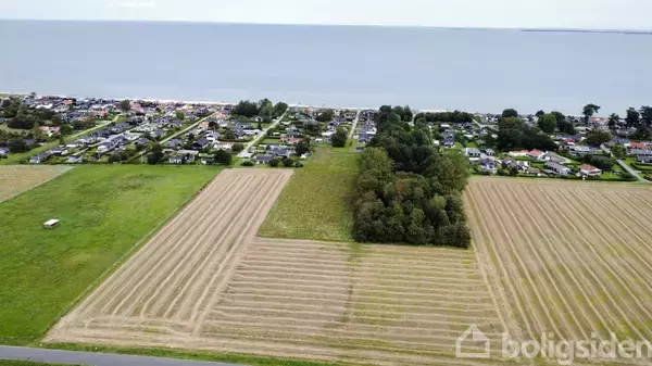 Landbrugsmarker ligger udbredt med stribede felter, omgivet af et boligområde i baggrunden og havudsigt i horisonten. En lille skov er midt på marken.