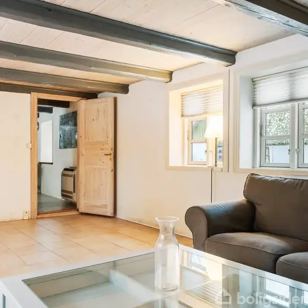 Et værelse med en grå sofa placeres i hjørnet og et glasbord foran. En åben trædør fører til et andet rum. Store vinduer giver naturligt lys.