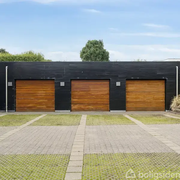 Tre garagedøre i trælook står lukkede på en sort bygning, omkranset af en brolagt indkørsel med græsstriber. Bagved ses buske og et enkelt træ under en blå himmel.