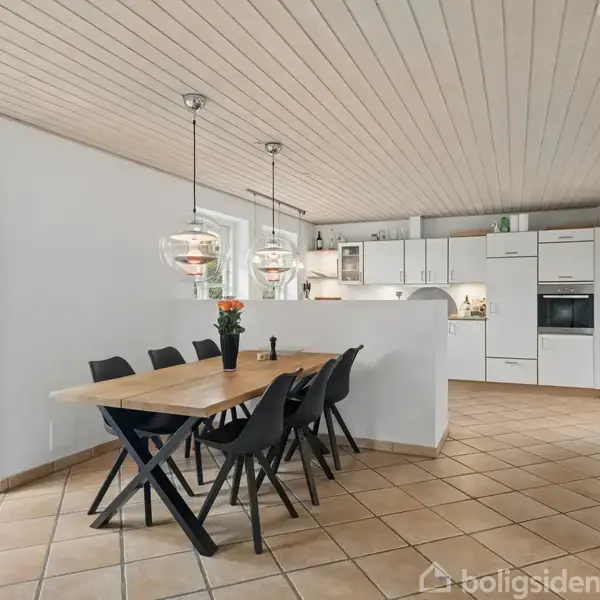 Et træbord med sorte stole står i en spisestue. Over bordet hænger moderne lamper. Baggrunden viser et hvidt køkken med skabe og en vægdekoreret verdenskort.