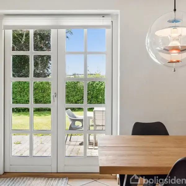 Dobbelt glasdør fører ud til terrasse med havemøbler og grøn have. Indendørs er et spisebord med sorte stole og en hængende lampe. En potteplante står på bordet.