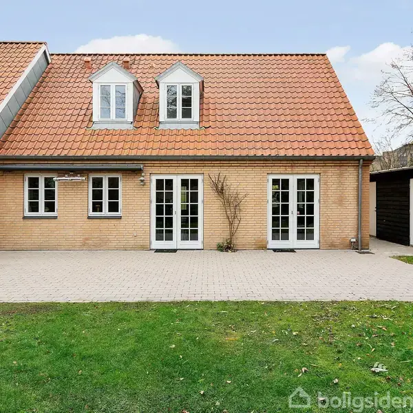 Hus med rødt tegltag og to kviste, ligger ved terrasse og græsplæne. Bygningen har to store glasdøre og er omgivet af træer og skur.