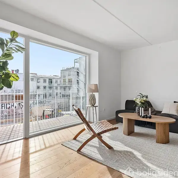 En lys stue indeholder en sort sofa, et træbord, og en lænestol på et tæppe. En stor plante står ved et vindue, der åbner ud til en balkon med byudsigt.