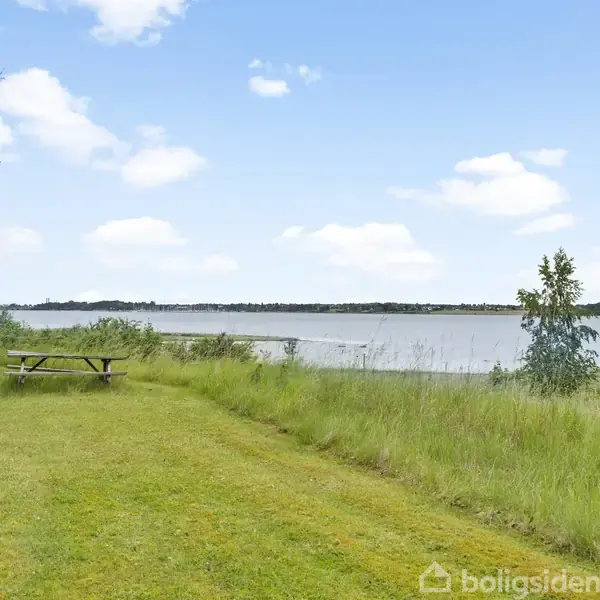 En træbænk og bord står på en græsplæne ved siden af en sø. Omgivet af træer og buske, ses en blå himmel med få skyer i baggrunden.