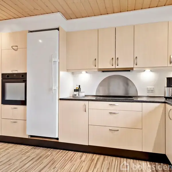 Køkken med lyse træskabe og indbygget ovn ved siden af et stort, hvidt køleskab. Bordpladen har en sort finish med en kaffemaskine placeret til højre. Loftet er træbeklædt.