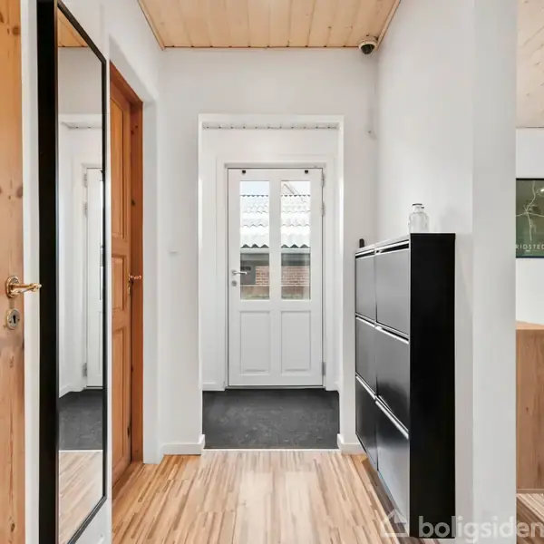 En entré med trægulv fører til en hvid hoveddør. Til højre står en sort skoreol, og til venstre er en spejlvæg og en trædør. En del af en spisestue ses.