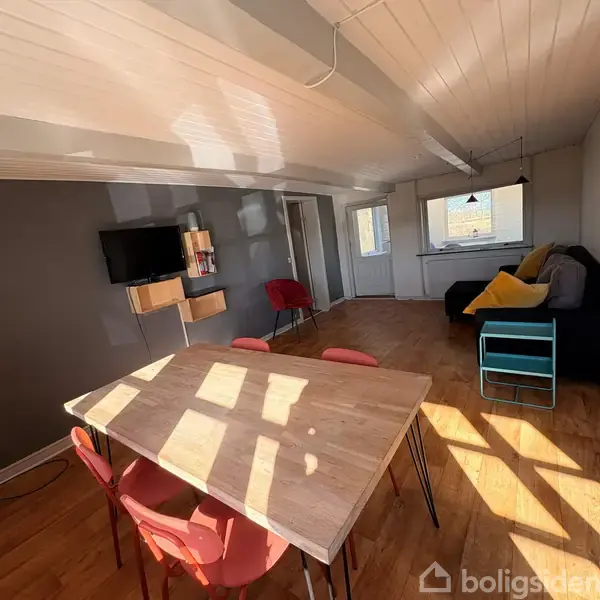 Stue med spisebord og fire røde stole i sollys. Fjernsynet hænger på en grå væg ved hylder. I baggrunden er en sofa med puder.