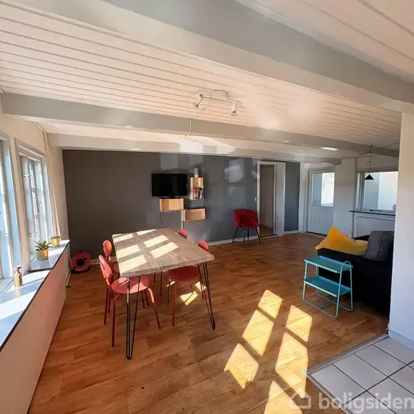 Spisebord med røde stole står i lyst rum med trægulv. Væggen har mørkegrå maling, en fladskærm og hylder. Et vindue lader sollys ind, belysende en gul pude på en sort sofa.
